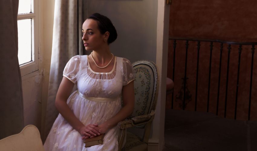 Woman in white Regency style empire waist dress sitting by a window in a historic interior