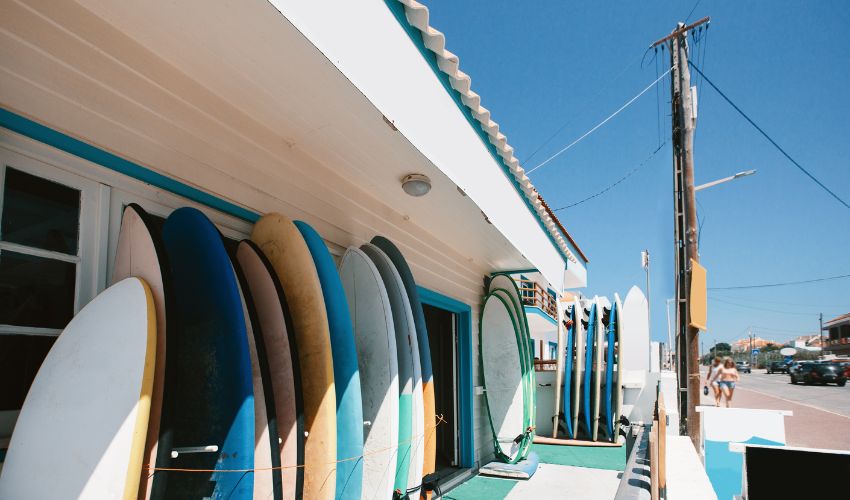Exterior of a traditional Portuguese surf shop featuring a row of colorful surfboards leaning against a white and teal building