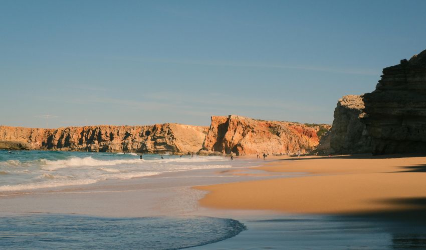 Wide golden sand beach with rocky cliffs and gentle ocean waves under a clear blue sky on the Alentejo Coast