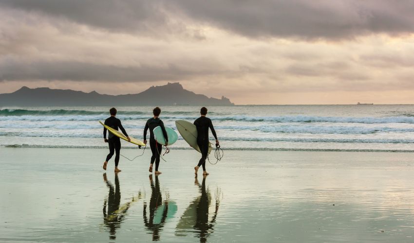 Three surfers in wetsuits carrying surfboards toward the ocean waves at sunset on a misty beach in Portugal