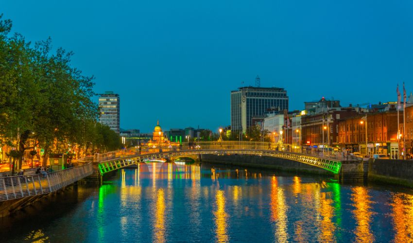 A lit-up pedestrian bridge over a calm river at dusk with city buildings reflecting in the water