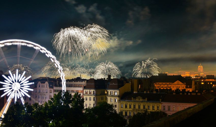 Fireworks lighting up the night sky over city buildings and a glowing Ferris wheel