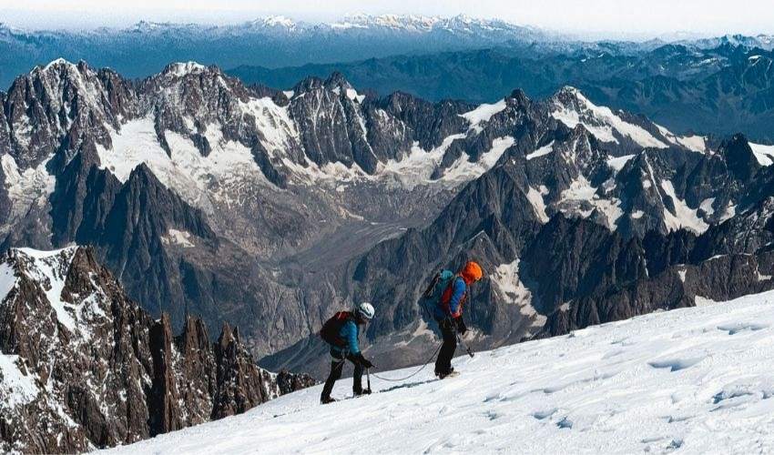 Two hikers with backpacks climbing a steep, snow-covered peak in the French Alps
